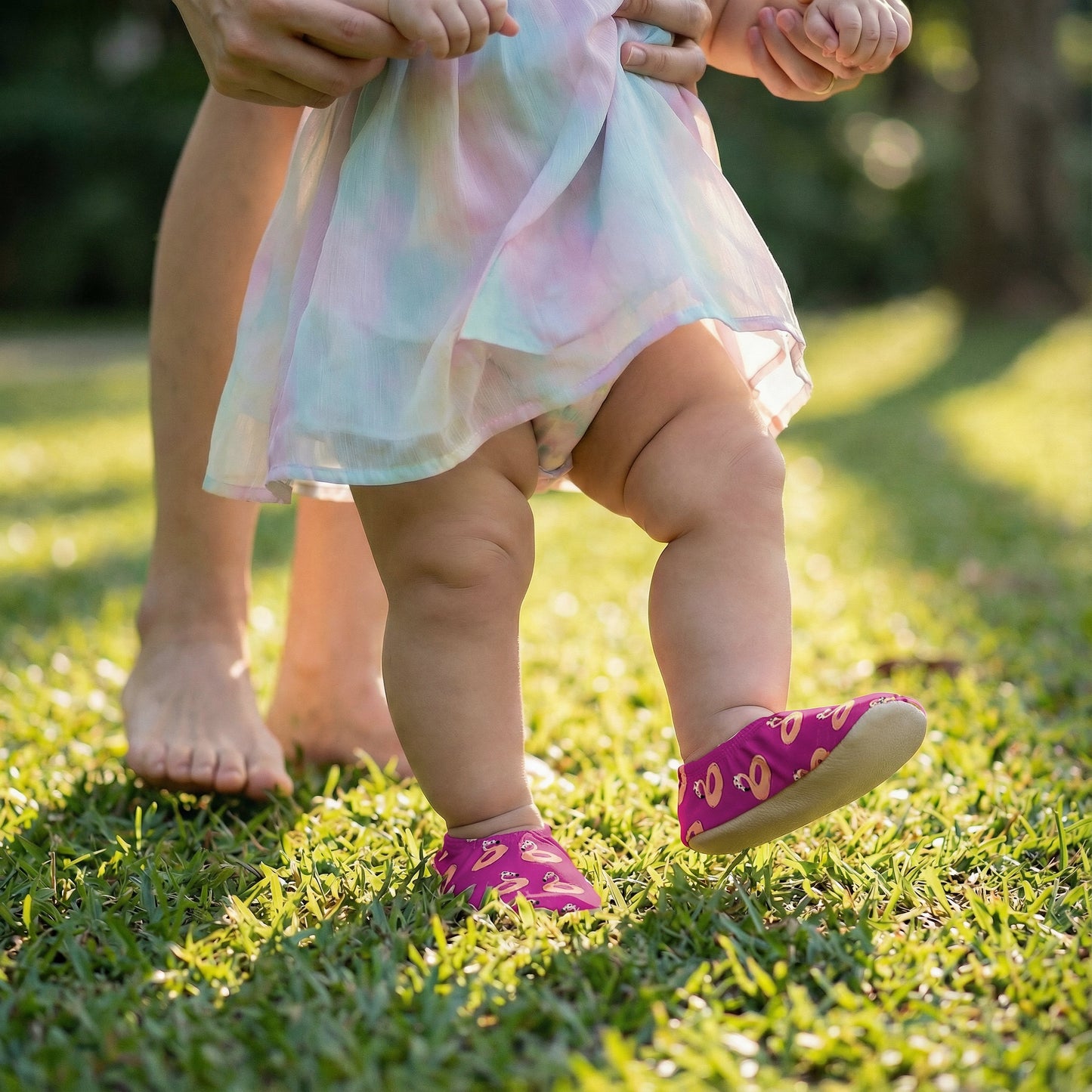 Baby in a colorful dress and non-slip socks standing on grass with adult's feet visible.
