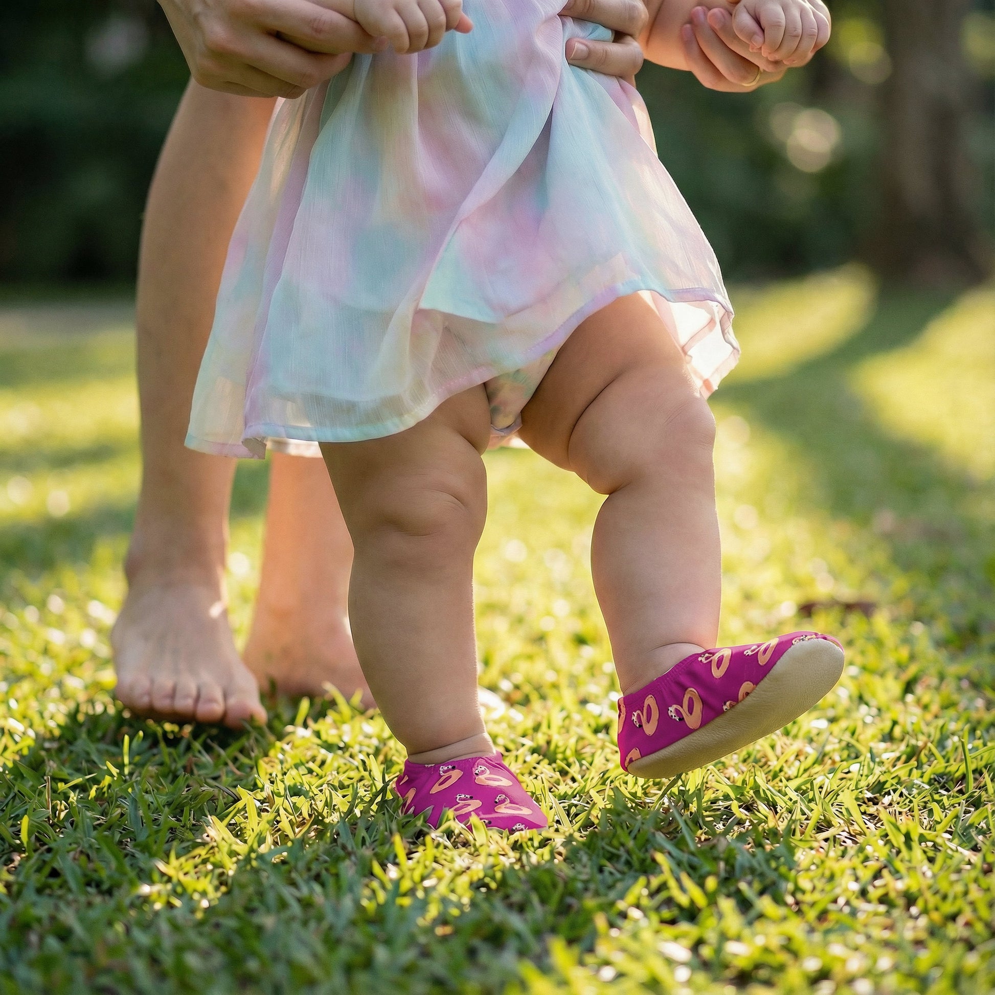 Baby in a colorful dress and non-slip socks standing on grass with adult's feet visible.